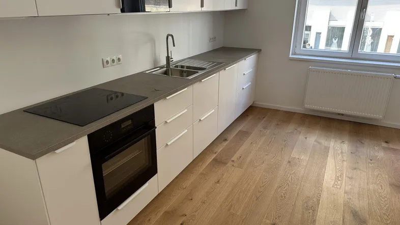Moderne Einbauküche mit weißen Schränken, dunkler Arbeitsplatte und Holzboden, gut beleuchtet durch ein Fenster.