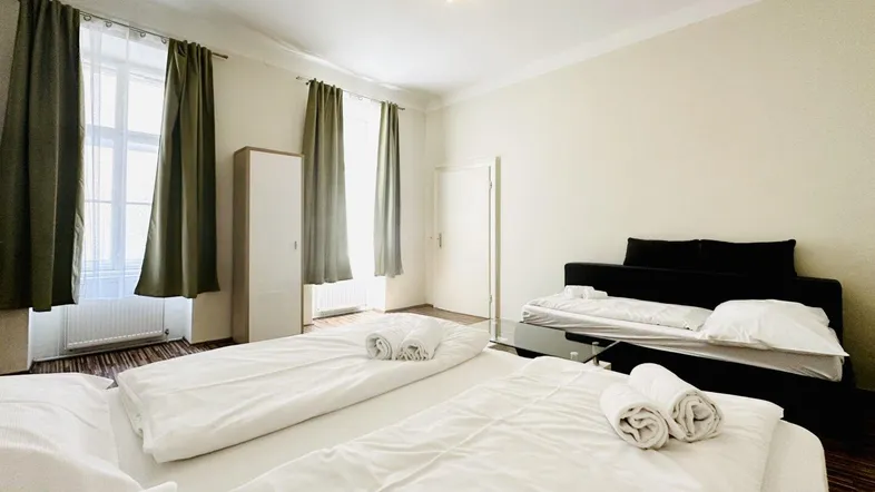 Helles Schlafzimmer mit zwei Betten, Parkettboden und hohen Fenstern mit Vorhängen.