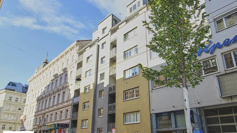 Moderne Wohngebäude-Fassade mit mehreren Balkonen und angrenzenden Altbauten in einer städtischen Umgebung.