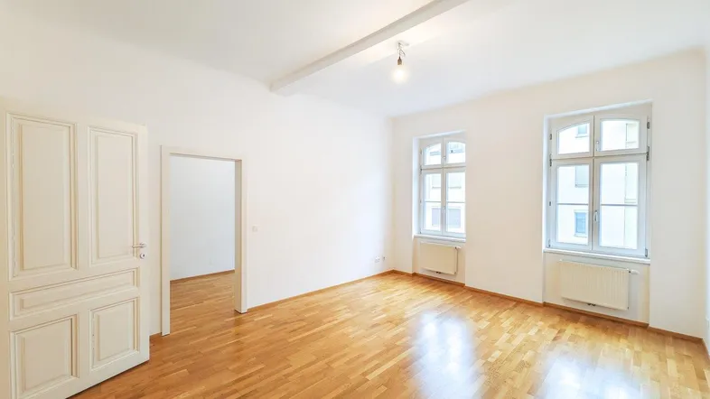 Helles Wohnzimmer mit Parkettboden, hohen Decken und zwei Fenstern.
