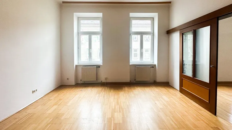 Heller Raum mit Parkettboden, zwei Fenstern und Schiebetür zu einem weiteren Zimmer.