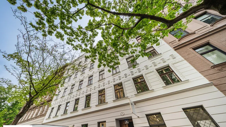 Klassische Altbau-Fassade mit vielen Fenstern und grünen Bäumen im Vordergrund unter blauem Himmel.
