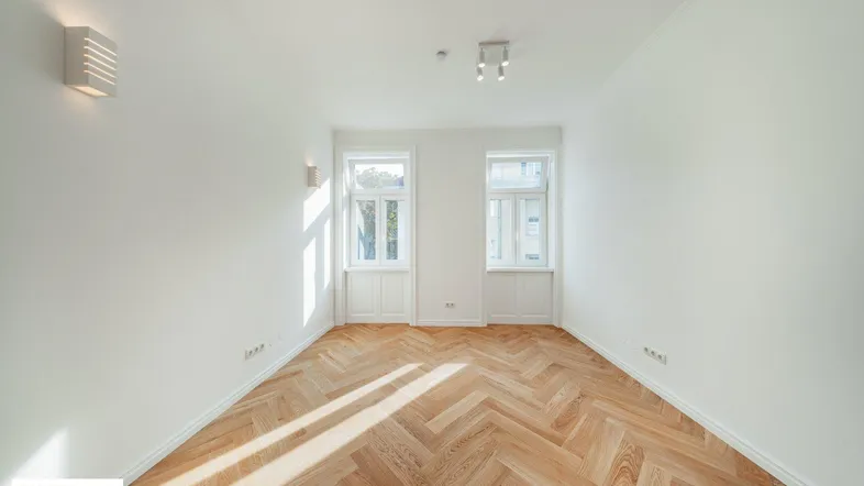 Helles Zimmer mit Fischgrätparkett und zwei Fenstern, das viel natürliches Licht hereinlässt.