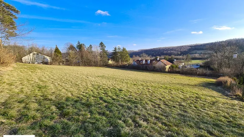 Weitläufige Grünfläche mit Blick auf umliegende Häuser und bewaldete Hügel unter blauem Himmel.
