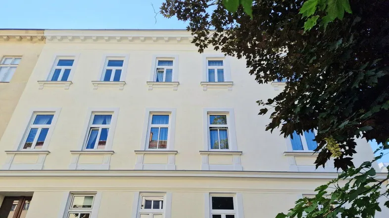 Klassische Fassade eines Mehrfamilienhauses mit vielen Fenstern und grünen Bäumen im Vordergrund.