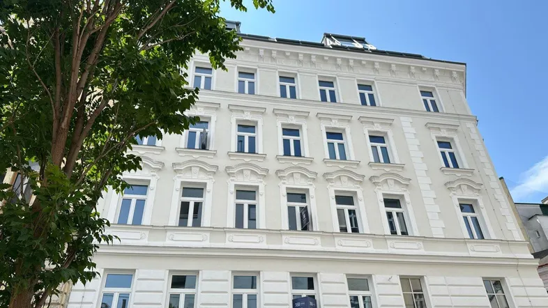 Helle, klassische Fassade eines Altbaus mit vielen Fenstern und grünen Bäumen im Vordergrund.