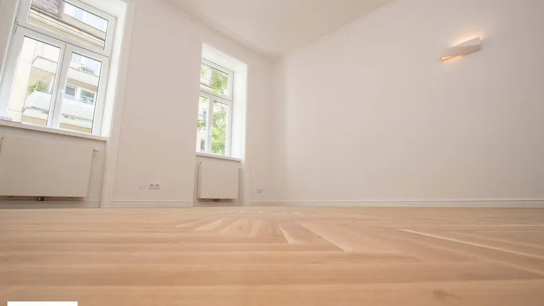 Helles Zimmer mit weiß gestrichenen Wänden und elegantem Fischgrätparkettboden, ideal für Wohn- oder Schlafbereich.