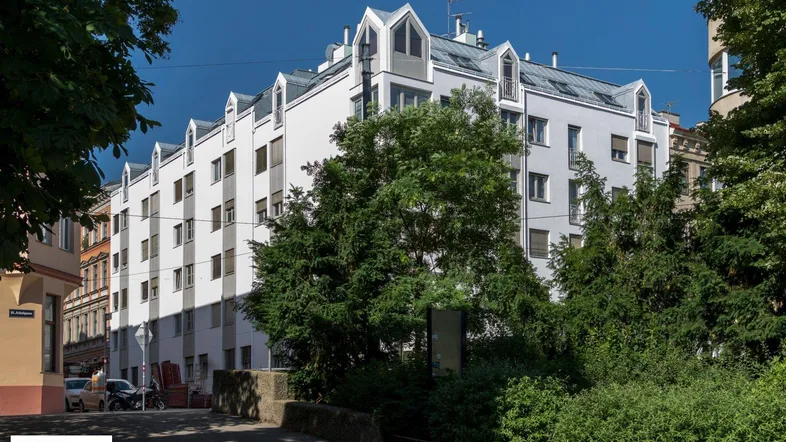 Weißes Mehrfamilienhaus mit markanter Dachform, Gaubenfenstern und vielen Bäumen im Vordergrund.