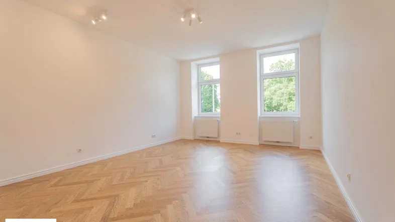 Helles, leeres Zimmer mit Fischgrätparkett und zwei großen Fenstern, die viel Tageslicht hereinlassen.