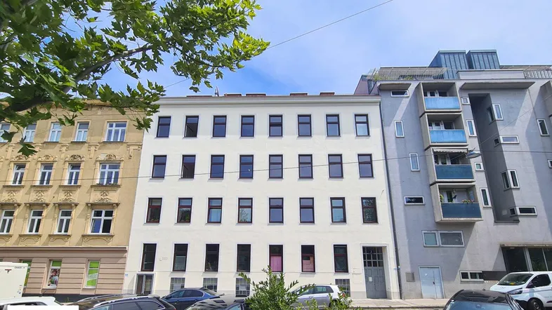 Weißes Mehrfamilienhaus mit vielen Fenstern an einer städtischen Straße, umgeben von anderen Gebäuden und parkenden Autos.