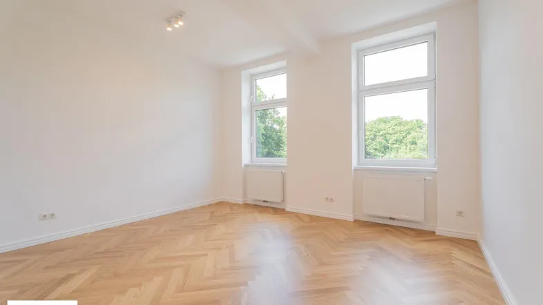 Helles Zimmer mit Fischgrätparkett und zwei Fenstern, die einen Blick ins Grüne bieten.