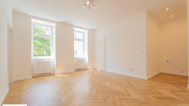Helles Zimmer mit Fischgrätparkettboden, weißen Wänden und zwei großen Fenstern, die viel Licht hereinlassen.