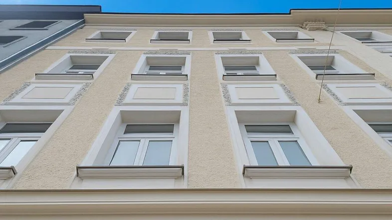 Helle Fassade eines Altbaus mit vielen Fenstern unter blauem Himmel an einem sonnigen Tag.