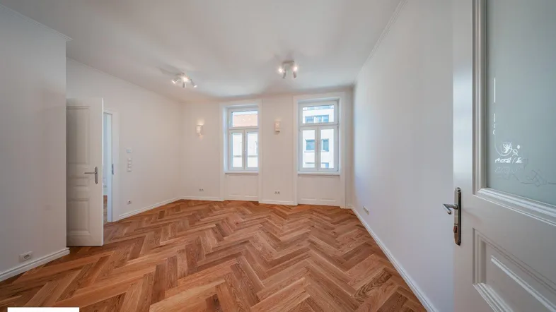 Geräumiges Zimmer mit Fischgrätparkett und zwei Fenstern, die viel Tageslicht hereinlassen.