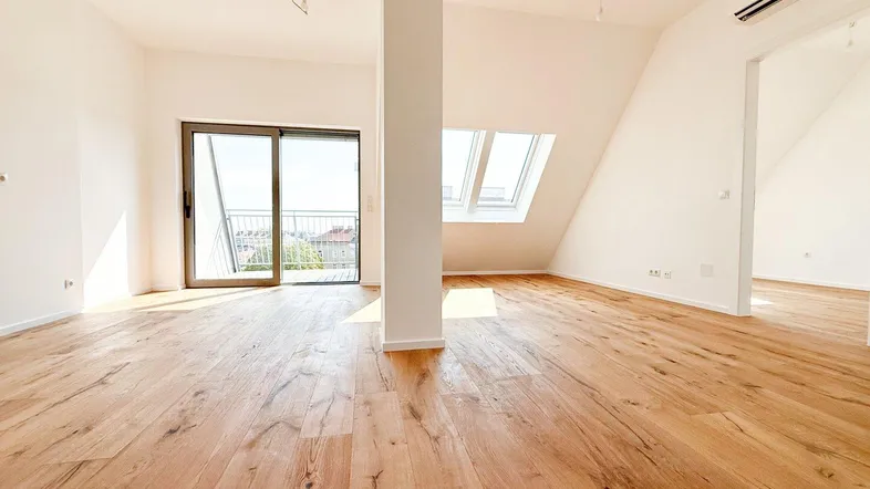 Heller, leerer Wohnbereich mit Holzboden, Dachschrägen, großen Fenstern und Zugang zum Balkon.