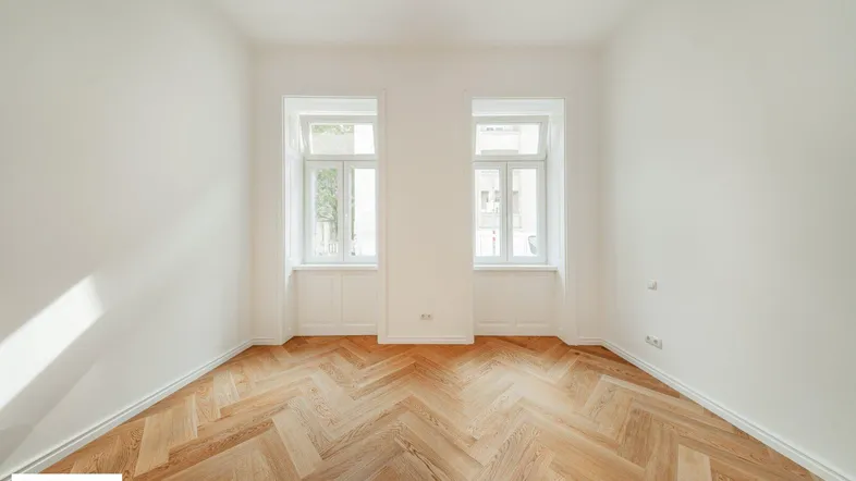 Heller Raum mit Fischgrätparkettboden und zwei Fenstern, frisch renoviert und einladend.