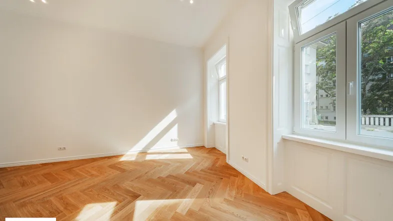 Heller Raum mit Fischgrätparkettboden und großen Fenstern, durch die Sonnenlicht einfällt.