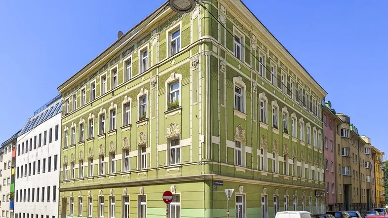Grünes Eckgebäude mit vielen Fenstern und klassischer Fassadenverzierung in städtischer Umgebung.