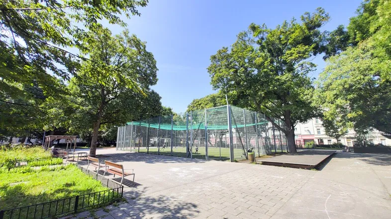 Grüner Parkbereich mit einem eingezäunten Sportplatz, umgeben von Bäumen und Sitzbänken an einem sonnigen Tag.