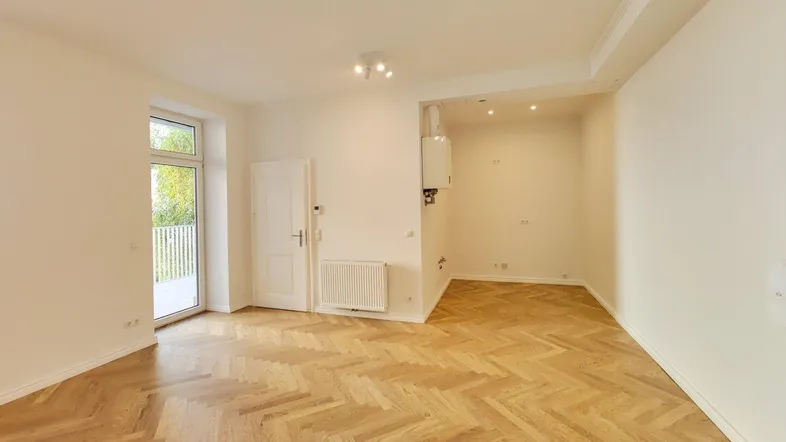 Helles, leeres Zimmer mit Fischgrätparkettboden, Heizkörper und Zugang zu einem Balkon.