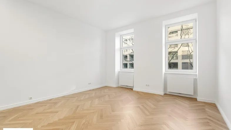 Helles Zimmer mit Fischgrätparkett und zwei großen Fenstern, ideal als Wohn- oder Schlafzimmer.