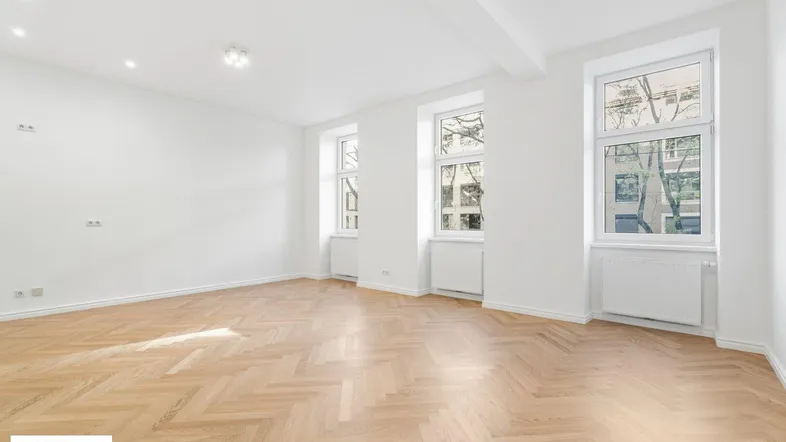Helles, leeres Zimmer mit Fischgrätparkett und großen Fenstern, ideal als Wohnbereich.