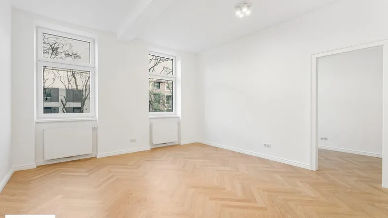 Helles Wohnzimmer mit Fischgrätparkett und zwei großen Fenstern mit Blick ins Grüne.