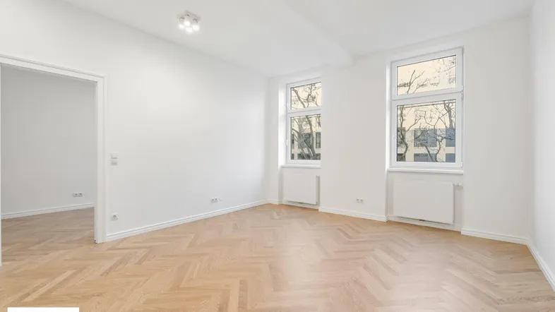 Helles, leeres Zimmer mit Fischgrätparkett und zwei Fenstern, bereit zur individuellen Gestaltung.