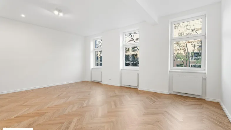 Helles, leeres Zimmer mit Fischgrätparkett und drei großen Fenstern, die viel Tageslicht hereinlassen.