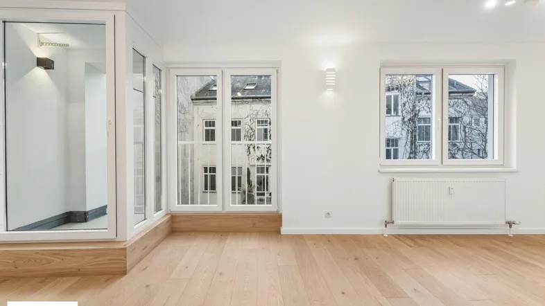 Helles Zimmer mit Holzboden und großen Fenstern, die einen Blick auf die umliegenden Gebäude bieten.