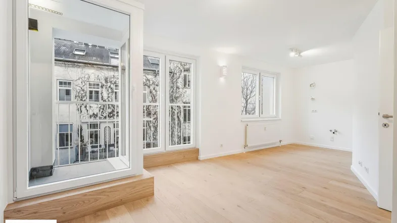 Helles, leeres Zimmer mit Holzboden, großen Fenstern und Zugang zu einem kleinen Balkon.