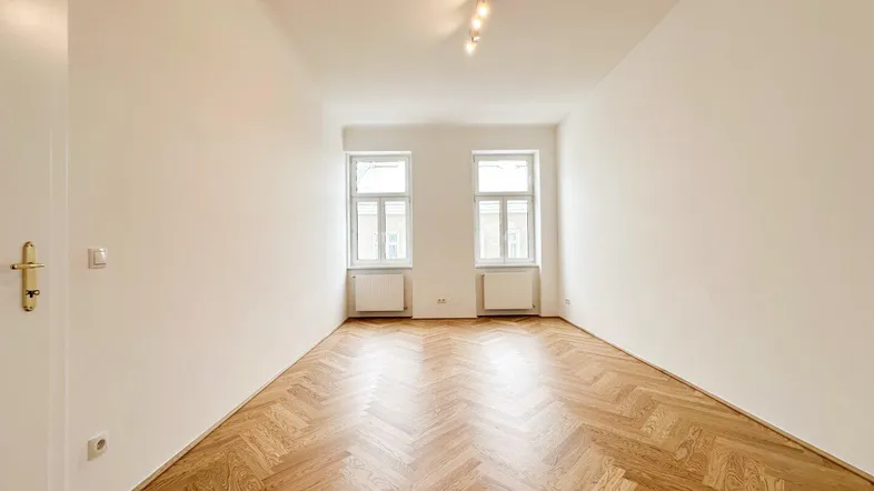 Heller Raum mit Fischgrätparkettboden und zwei Fenstern, ideal als Wohnzimmer nutzbar.