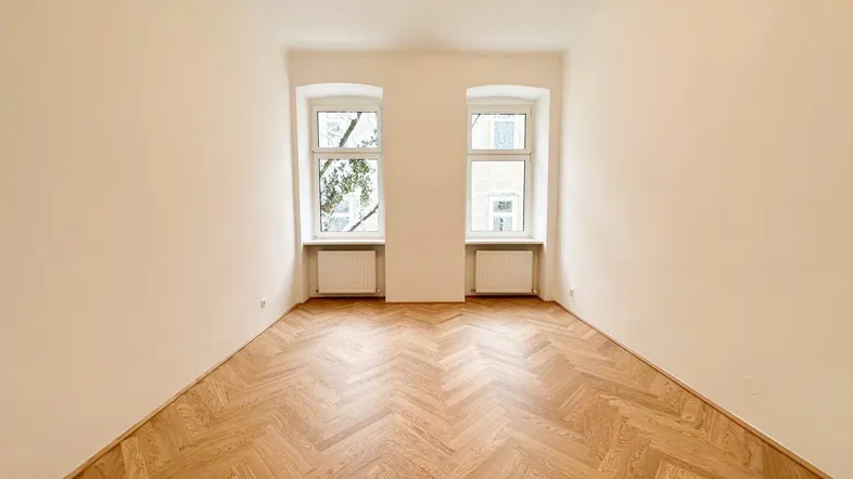 Helles Zimmer mit Fischgrätparkett und zwei Fenstern, ideal als Wohn- oder Schlafzimmer nutzbar.