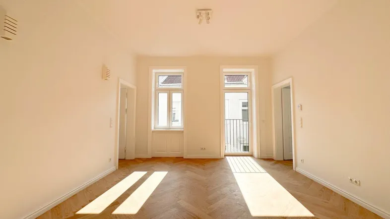 Helles Zimmer mit Fischgrätparkettboden, hohen Decken und Zugang zu einem französischen Balkon.
