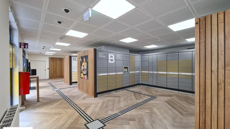Moderner Innenraum einer Poststation mit Paketfächern, Holzverkleidung und einem Fischgrätmuster auf dem Boden.