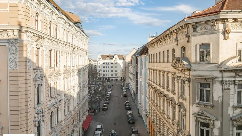 Blick auf eine belebte Stadtstraße mit historischen Gebäuden und parkenden Autos unter blauem Himmel.