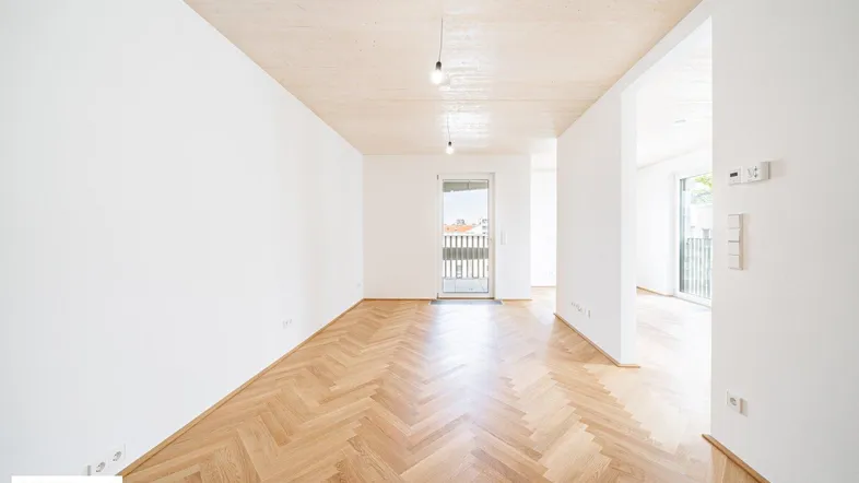 Helles, leeres Zimmer mit Fischgrätparkettboden und Zugang zu einem französischen Balkon.