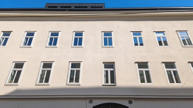 Helle Fassade eines Mehrfamilienhauses mit vielen Fenstern unter blauem Himmel.