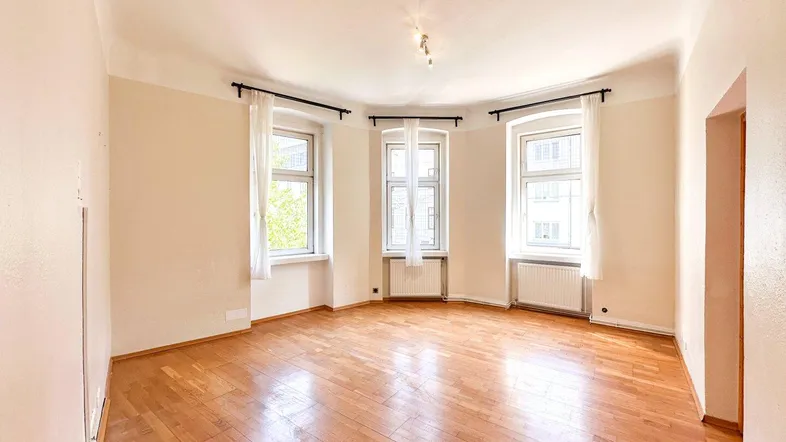 Helles Wohnzimmer mit Parkettboden und drei großen Fenstern, die viel Tageslicht hereinlassen.