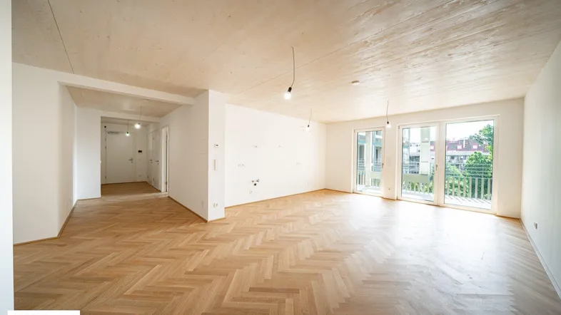 Helles, leeres Wohnzimmer mit Fischgrätparkett, Holzbalkendecke und Zugang zu einem Balkon.