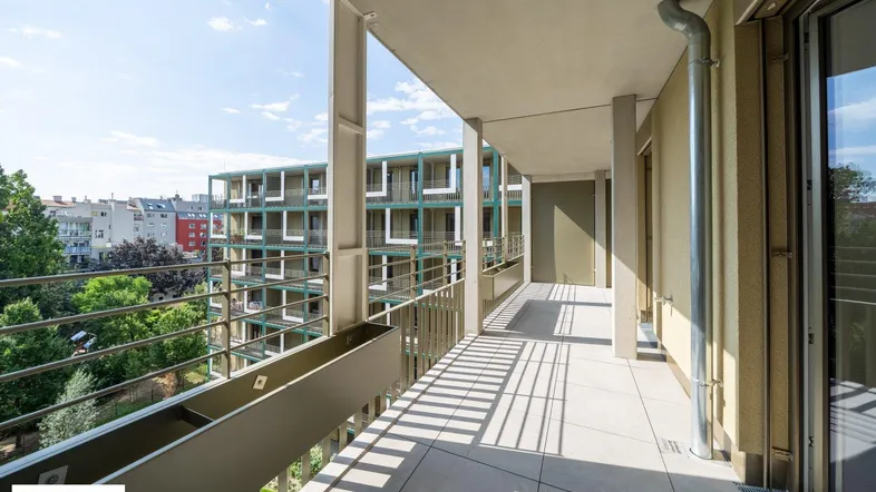 Langer Balkon mit Fliesenboden und Blick auf moderne Gebäude sowie grüne Bäume.