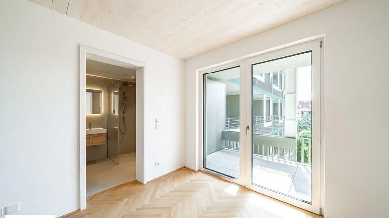 Helles Zimmer mit Fischgrätparkett, Holzbalkendecke und Zugang zu einem Balkon, daneben ein modernes Badezimmer.