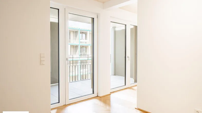 Heller Innenraum mit Fischgrätparkett und großen Fenstern, die Zugang zu einem Balkon bieten.