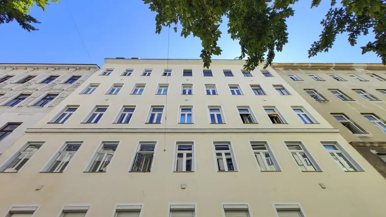 Die helle Fassade des Mehrfamilienhauses mit vielen Fenstern unter blauem Himmel.