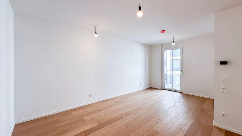 Helles, leeres Zimmer mit Holzboden und Zugang zu einem Balkon, ideal für ein Wohnzimmer.