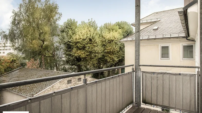 Balkon mit Holzboden und Blick auf grüne Bäume sowie umliegende Gebäude.
