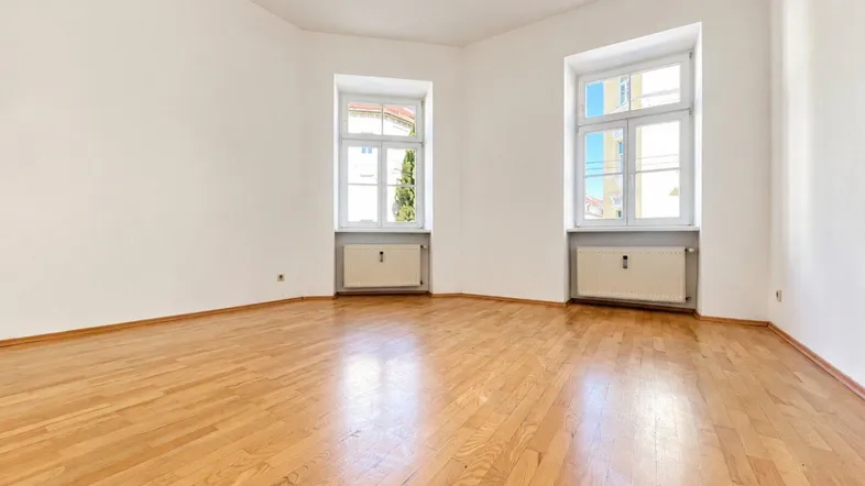 Helles, leeres Zimmer mit Holzboden und zwei Fenstern, bereit zur individuellen Gestaltung.