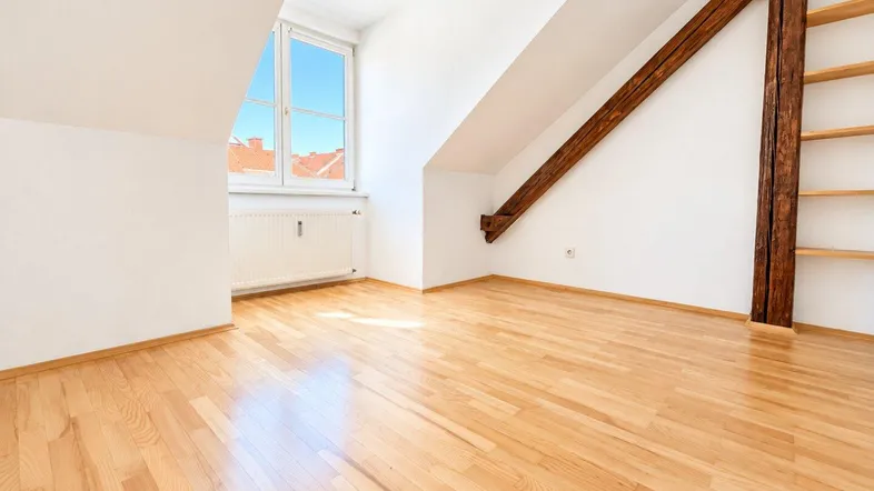 Lichtdurchflutetes Dachzimmer mit Holzboden, sichtbaren Holzbalken und einem großen Fenster.