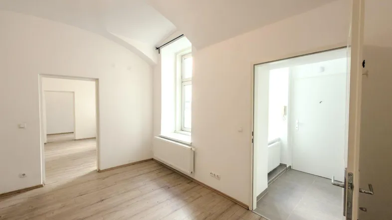 Helles Zimmer mit Holzboden, weiß gestrichenen Wänden und einem Fenster, das viel Tageslicht hereinlässt.