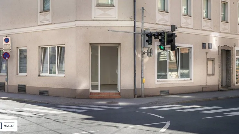 Gebäude an einer Straßenecke mit Schaufenstern und einem Eingang im Erdgeschoss, beleuchtet von Sonnenlicht.